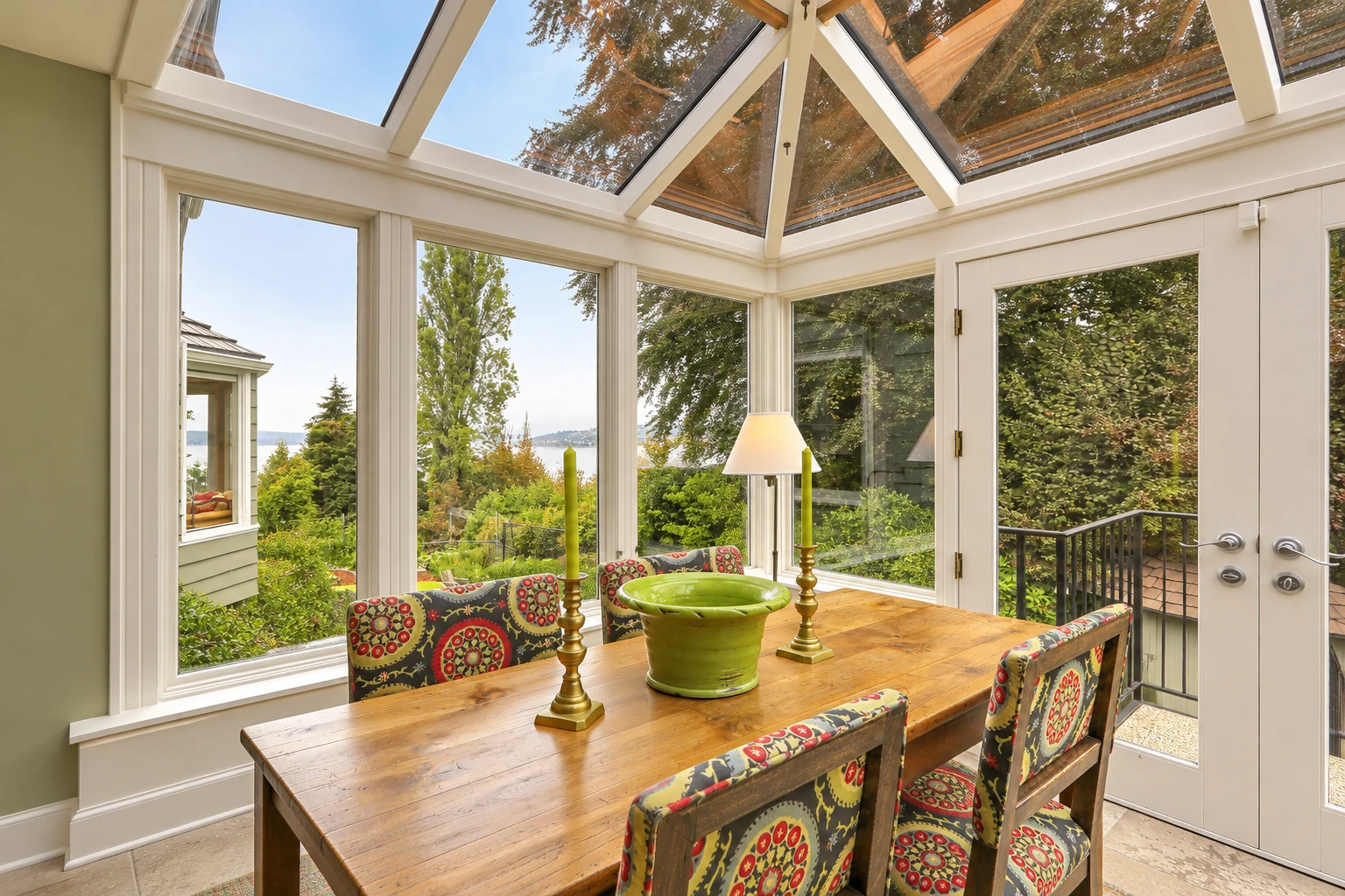 A custom sunroom with a glass-paneled peaked ceiling contains a wooden dining table and patterned chairs, with garden views through wide windows.