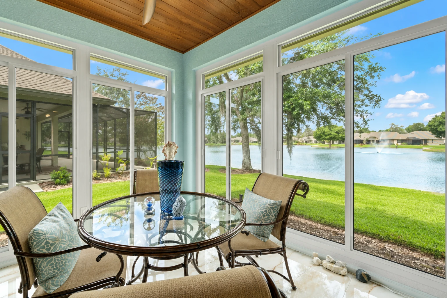 Enclosed patio room with white-framed windows, a glass table and cushioned chairs overlooking a lake.