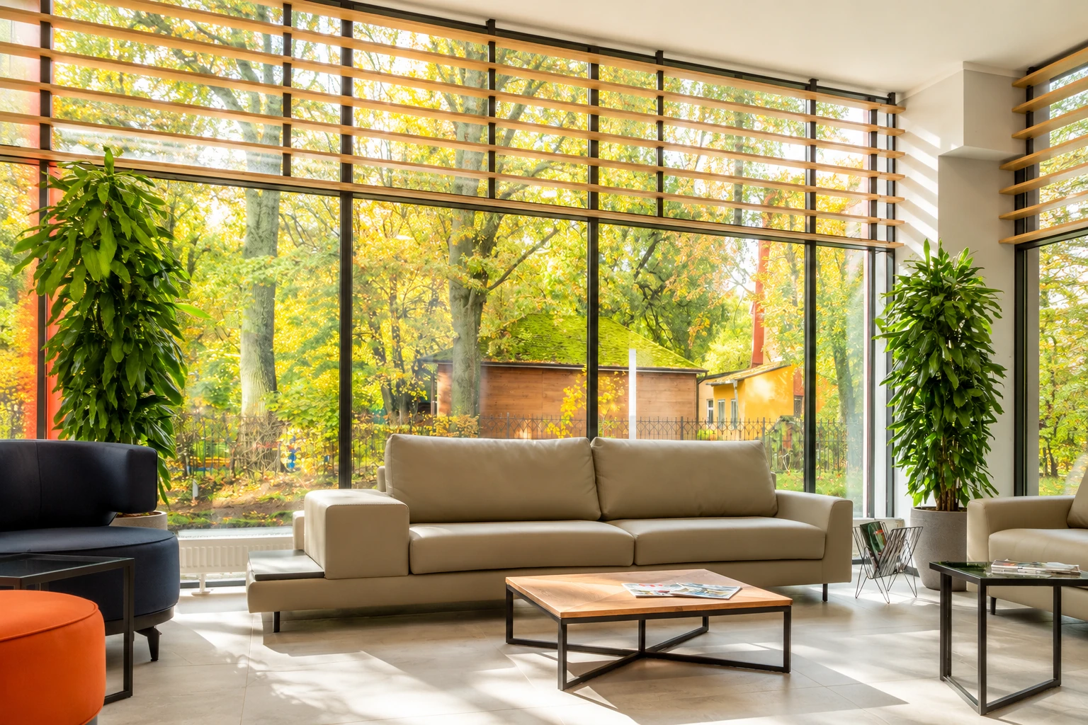 Bright sunroom living area with a beige sofa, plants, and broad windows shaded by horizontal wooden slats.