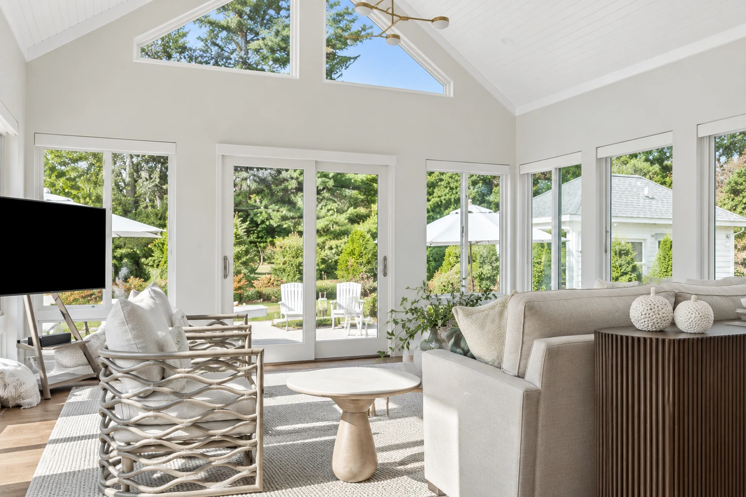 Bright sunroom with a vaulted white ceiling, large windows, and a neutral seating area overlooking a garden.